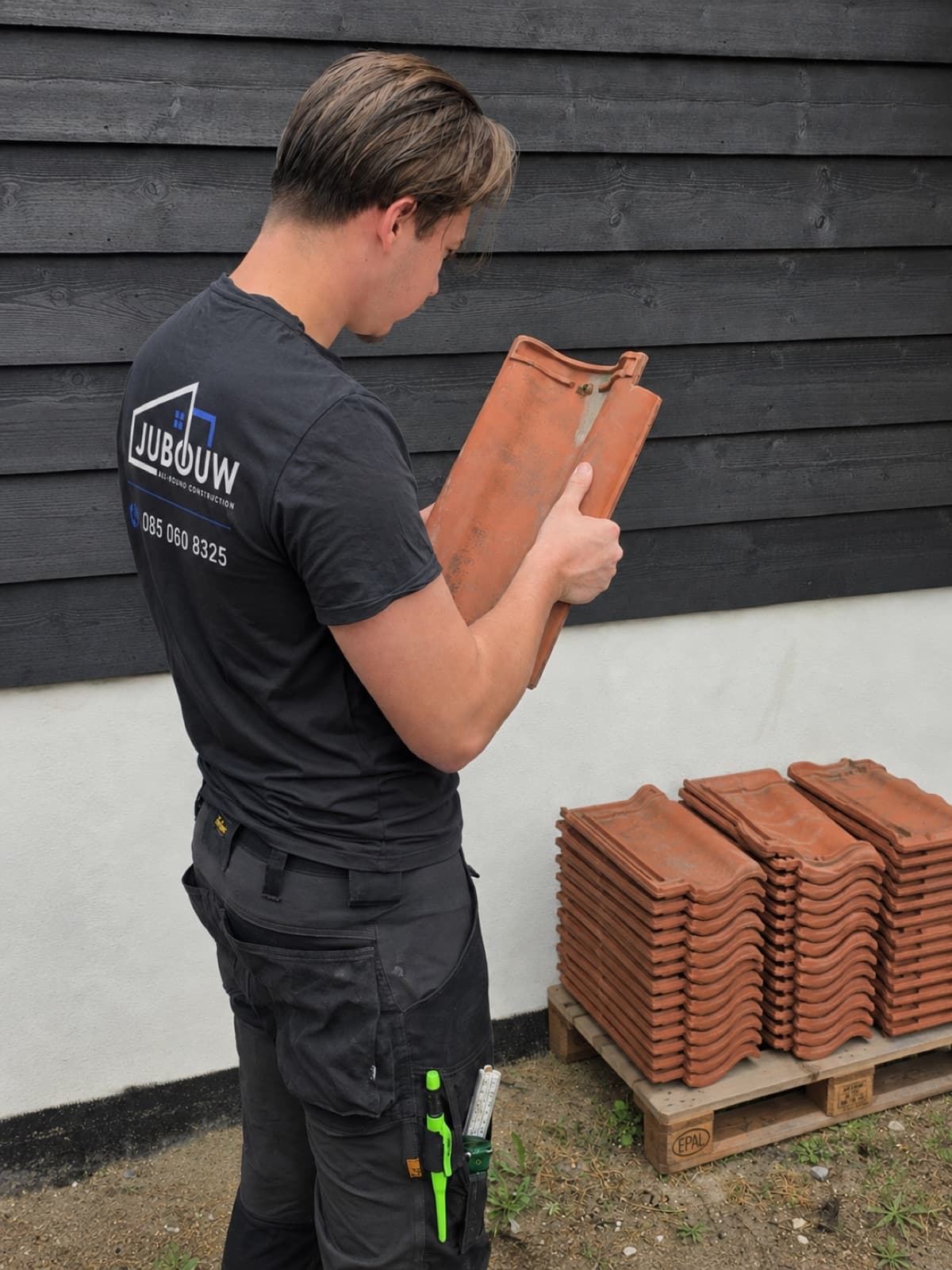 Monteur van Jubouw met een dakpan naast een pallet nieuwe dakpannen